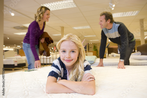 Photo: Girl (6-8) on bed in furniture shop, parents in background, smiling, portrait Girl (6-8) on bed in furniture shop, parents in background, smiling, portrait