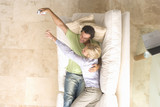 Photo: Young couple embracing on sofa, woman reaching for remote control from man, elevated view Young couple embracing on sofa, woman reaching for remote control from man, elevated view