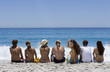 Group of teenagers (17-19) sitting in line on beach, looking at horizon over sea, rear view