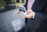 Photo: Businessman standing in lobby with luggage, using mobile phone, mid-section (differential focus) Businessman standing in lobby with luggage, using mobile phone, mid-section (differential focus)
