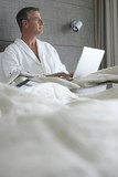 Photo: Businessman in bathrobe sitting in hotel bed, using laptop, thinking, surface level Businessman in bathrobe sitting in hotel bed, using laptop, thinking, surface level