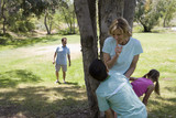 Photo: Family playing hide and seek in park, mother and children (8-11) hiding behind tree, father looking Family playing hide and seek in park, mother and children (8-11) hiding behind tree, father looking