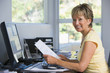 Woman in home office with computer and paperwork smiling
