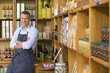 Photo: Male grocery shop owner in apron standing beside shelf display, arms folded, smiling, portrait Male grocery shop owner in apron standing beside shelf display, arms folded, smiling, portrait