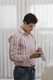 Photo: Businessman standing beside desk in office, using personal electronic organiser, side view Businessman standing beside desk in office, using personal electronic organiser, side view