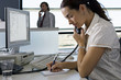 Photo: Businesswoman working at desk in office, using telephone, writing in notepad, man in background Businesswoman working at desk in office, using telephone, writing in notepad, man in background