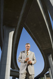 Photo: Businessman using electronic organiser beneath overpasses, portrait, low angle view Businessman using electronic organiser beneath overpasses, portrait, low angle view