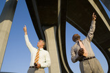 Photo: Two businessmen with mobile phones beneath overpasses, pointing upwards, low angle view Two businessmen with mobile phones beneath overpasses, pointing upwards, low angle view
