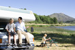Photo: Man smiling at son (10-12) on bonnet of motor home by lake, mother reading in background Man smiling at son (10-12) on bonnet of motor home by lake, mother reading in background
