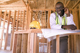 Photo: Businessman with blueprints using mobile phone in partially built house, smiling, portrait Businessman with blueprints using mobile phone in partially built house, smiling, portrait