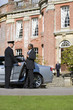 Photo: Chauffeur holding open door of car for businessman by manor house, low angle view Chauffeur holding open door of car for businessman by manor house, low angle view
