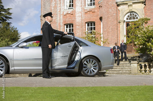Photo: Chauffeur holding open door of car by manor house, businessmen in background Chauffeur holding open door of car by manor house, businessmen in background