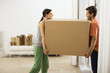 Photo: Couple moving house, carrying large cardboard box in sparse room, face to face, smiling, profile Couple moving house, carrying large cardboard box in sparse room, face to face, smiling, profile