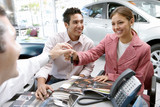 Photo: Young couple sitting at desk in car showroom, salesman handing key to woman, smiling Young couple sitting at desk in car showroom, salesman handing key to woman, smiling