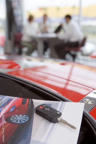 Photo: Salesman talking to customers in car showroom, focus on car key and photograph in foreground Salesman talking to customers in car showroom, focus on car key and photograph in foreground