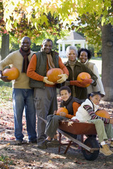 Photo: Multi-generational family standing in autumn garden, holding pumpkins, children (7-9) in wheelbarrow, portrait Multi-generational family standing in autumn garden, holding pumpkins, children (7-9) in wheelbarrow, portrait