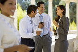 Photo: Small group of business colleagues standing in colonnade, holding paper cups, talking, side view Small group of business colleagues standing in colonnade, holding paper cups, talking, side view