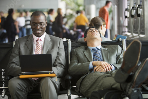Photo: Two businessmen sitting in airport terminal, one man using laptop, other man resting with feet up on luggage Two businessmen sitting in airport terminal, one man using laptop, other man resting with feet up on luggage