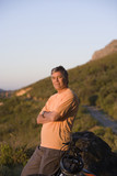 Photo: Mature man taking break from cycling on mountain trail, standing beside rock, arms folded, looking at scenery Mature man taking break from cycling on mountain trail, standing beside rock, arms folded, looking at scenery
