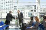 Photo: Two businessmen shaking hands beside large window in airport departure lounge, other passengers waiting in seats, side view Two businessmen shaking hands beside large window in airport departure lounge, other passengers waiting in seats, side view