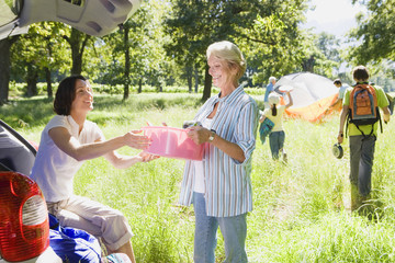 Photo: Multi-generational family unloading car on camping trip, adult daughter passing mother container from car boot Multi-generational family unloading car on camping trip, adult daughter passing mother container from car boot