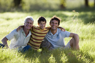 Photo: Boy (8-10) sitting with arms around father and grandfather in field, smiling, front view, portrait Boy (8-10) sitting with arms around father and grandfather in field, smiling, front view, portrait