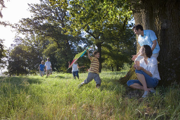 Photo: Multi-generational family exploring woodland clearing, parents resting beside tree, boy (8-10) playing with kite Multi-generational family exploring woodland clearing, parents resting beside tree, boy (8-10) playing with kite