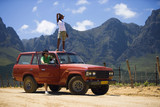 Photo: Young couple standing beside parked red SUV on dirt track in mountain valley, woman on roof, looking at scenery Young couple standing beside parked red SUV on dirt track in mountain valley, woman on roof, looking at scenery