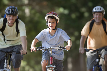 Photo: Boy (8-10), in cycling helmet, mountain biking on woodland trail with father and grandfather, front view Boy (8-10), in cycling helmet, mountain biking on woodland trail with father and grandfather, front view