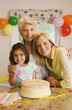 Photo: Girl (4-6) sitting with mother and grandmother at table beside birthday cake, making ‘thumbs up’ sign, smiling, portrait Girl (4-6) sitting with mother and grandmother at table beside birthday cake, making ‘thumbs up’ sign, smiling, portrait