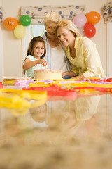 Photo: Girl (4-6) sitting at table beside birthday cake, mother and grandmother looking on, smiling, portrait (surface level) Girl (4-6) sitting at table beside birthday cake, mother and grandmother looking on, smiling, portrait (surface level)