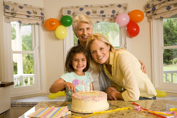 Photo: Girl (4-6) sitting with mother and grandmother at table beside birthday cake, smiling, portrait Girl (4-6) sitting with mother and grandmother at table beside birthday cake, smiling, portrait