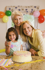 Photo: Girl (4-6) sitting with mother and grandmother at table beside birthday cake, making ‘thumbs up’ sign, smiling, portrait Girl (4-6) sitting with mother and grandmother at table beside birthday cake, making ‘thumbs up’ sign, smiling, portrait