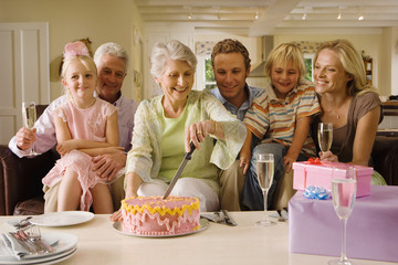 Photo: Three generation family sitting on sofa at home, senior woman cutting birthday cake, smiling Three generation family sitting on sofa at home, senior woman cutting birthday cake, smiling