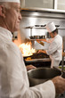 Photo: Chef frying food in flaming pan on gas hob in commercial kitchen, second chef carrying large pot, side view Chef frying food in flaming pan on gas hob in commercial kitchen, second chef carrying large pot, side view