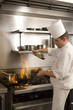 Photo: Chef pouring cooking oil into flaming pan on gas hob in commercial kitchen, side view Chef pouring cooking oil into flaming pan on gas hob in commercial kitchen, side view