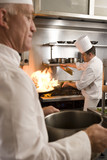 Photo: Chef frying food in flaming pan on gas hob in commercial kitchen, second chef carrying large pot, side view Chef frying food in flaming pan on gas hob in commercial kitchen, second chef carrying large pot, side view