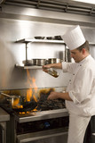 Photo: Chef pouring cooking oil into flaming pan on gas hob in commercial kitchen, side view Chef pouring cooking oil into flaming pan on gas hob in commercial kitchen, side view