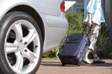 Photo: Senior woman pulling suitcase on wheels from parked car boot on driveway, rear view (surface level) Senior woman pulling suitcase on wheels from parked car boot on driveway, rear view (surface level)