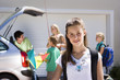 Photo: Family loading camping equipment into parked car boot on driveway, focus on girl (11-13) standing in foreground, smiling, portrait Family loading camping equipment into parked car boot on driveway, focus on girl (11-13) standing in foreground, smiling, portrait