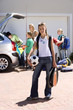 Photo: Family loading camping equipment into parked car boot on driveway, girl (11-13) carrying rucksack, soccer ball and tennis racquets in foreground, smiling, portrait Family loading camping equipment into parked car boot on driveway, girl (11-13) carrying rucksack, soccer ball and tennis racquets in foreground, smiling, portrait