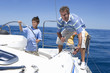Photo: Father and son (8-10) standing on deck of sailing boat out to sea, boy steering, man winding rope pulley of boat rigging, smiling Father and son (8-10) standing on deck of sailing boat out to sea, boy steering, man winding rope pulley of boat rigging, smiling