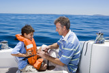 Photo: Father and son (8-10) sitting on deck of sailing boat out at sea, boy wearing orange life jacket, man tying strap, smiling, side view Father and son (8-10) sitting on deck of sailing boat out at sea, boy wearing orange life jacket, man tying strap, smiling, side view