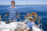 Photo: Family relaxing on deck of sailing boat out at sea, father standing at helm, steering, smiling, portrait Family relaxing on deck of sailing boat out at sea, father standing at helm, steering, smiling, portrait