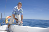 Photo: Man standing on deck of sailing boat out to sea, winding rope pulley of boat rigging, smiling (tilt) Man standing on deck of sailing boat out to sea, winding rope pulley of boat rigging, smiling (tilt)