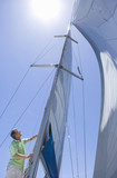 Photo: Man standing on deck of sailing boat out to sea, adjusting sail mast rigging, side view, low angle view (lens flare) Man standing on deck of sailing boat out to sea, adjusting sail mast rigging, side view, low angle view (lens flare)
