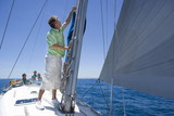 Photo: Family sitting on deck of sailing boat out to sea, watching father adjusting sail mast rigging in foreground, side view Family sitting on deck of sailing boat out to sea, watching father adjusting sail mast rigging in foreground, side view