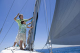 Photo: Father and son (8-10) standing on deck of sailing boat out to sea, adjusting sail mast rigging, side view Father and son (8-10) standing on deck of sailing boat out to sea, adjusting sail mast rigging, side view