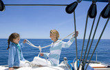 Photo: Mother and daughter (8-10) sitting at stern of sailing boat out at sea, tying knots in rope, smiling, profile Mother and daughter (8-10) sitting at stern of sailing boat out at sea, tying knots in rope, smiling, profile