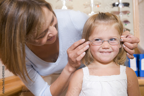 Woman trying eyeglasses on young girl at optometrists smiling